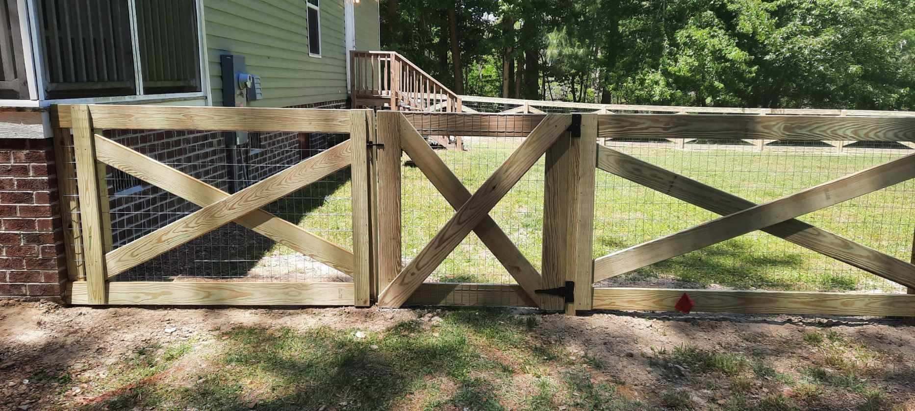 Wooden backyard gate with X-brace panels beside a house, set in a sunny yard with trees.