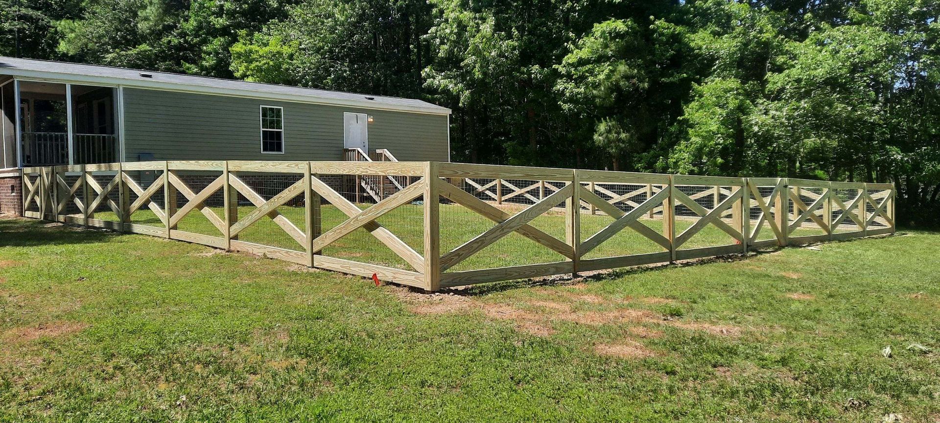 Wooden fenced enclosure beside a mobile home in a grassy yard with trees in the background