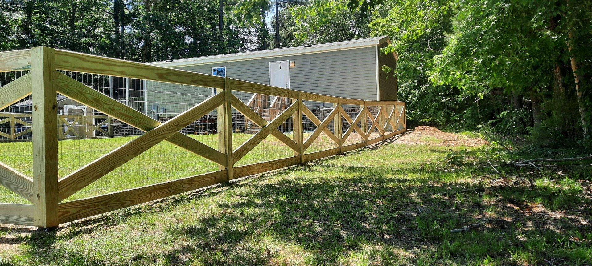 Wooden fence with X-bracing along a grassy path beside a white building in a wooded area
