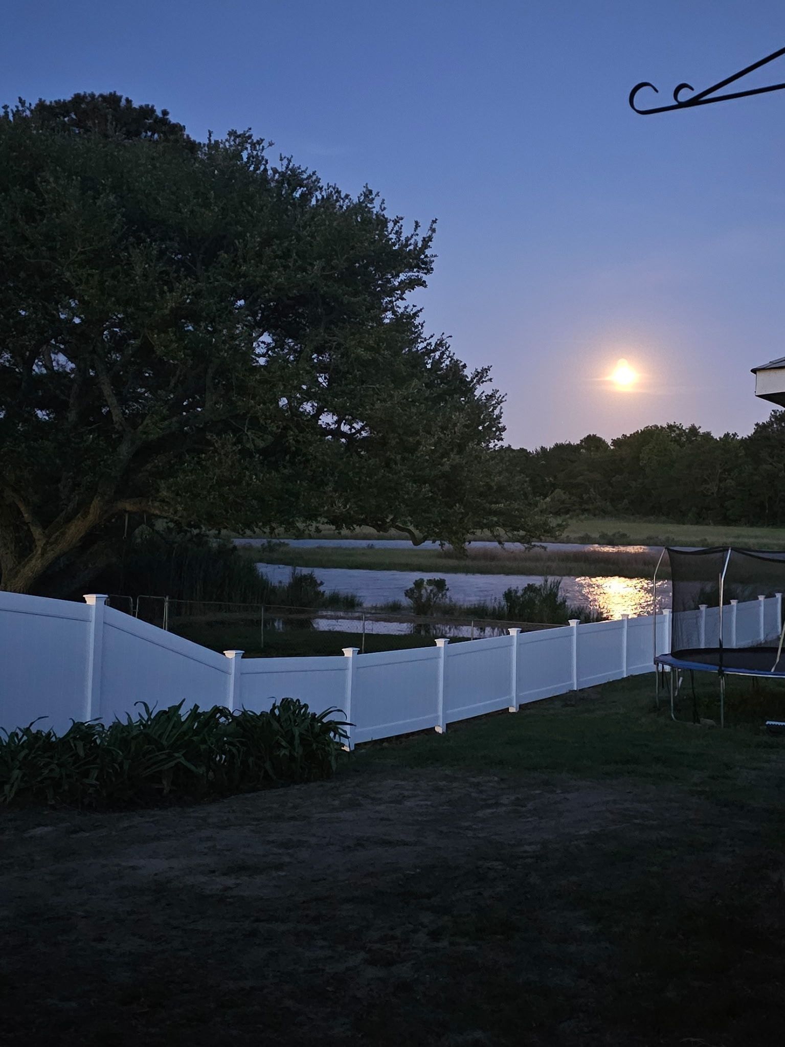 Moonrise over a river behind a white fence at dusk, with trees silhouetted against a blue sky