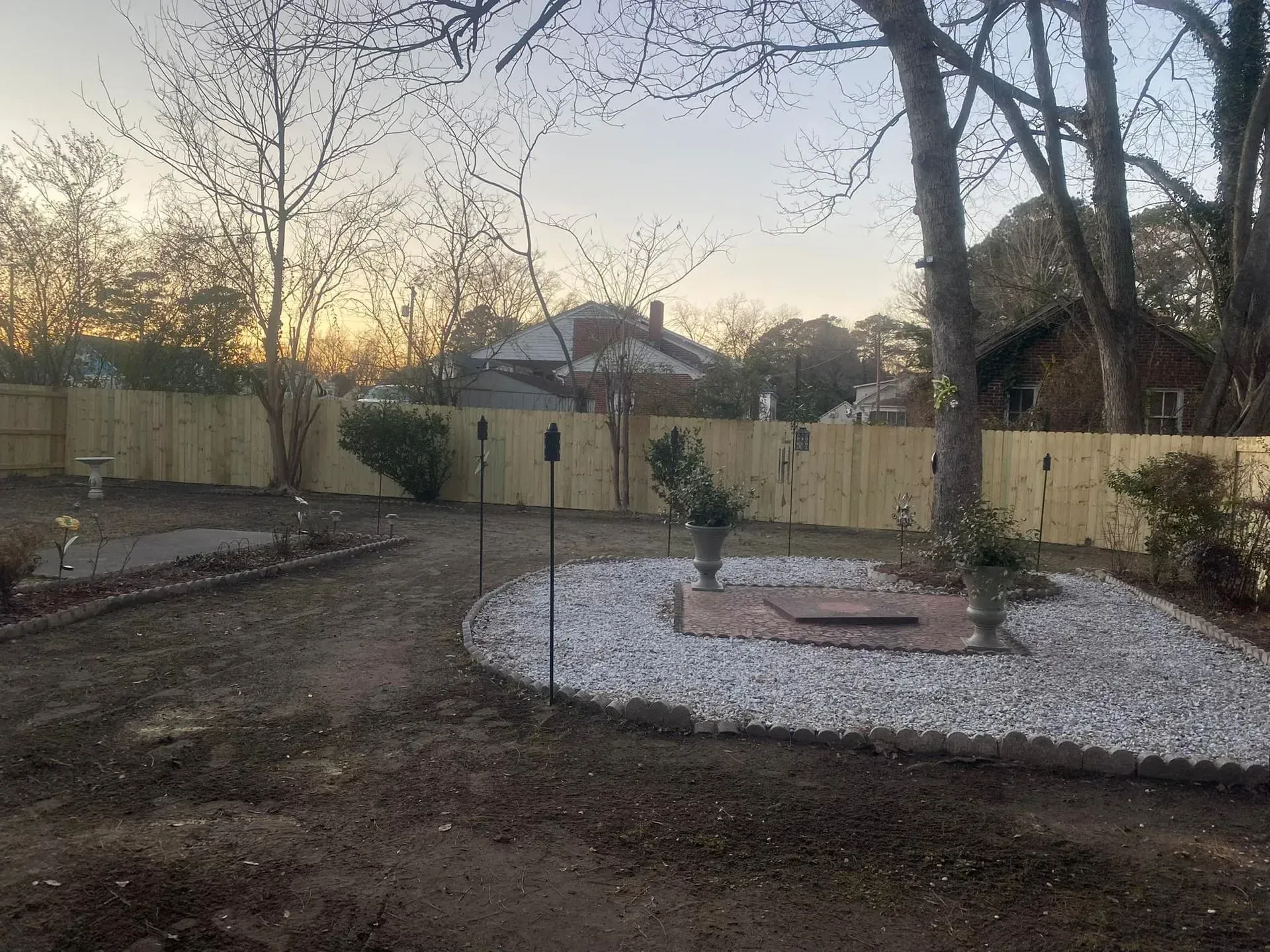 Backyard with wooden fence, fire pit surrounded by white stones, and bare trees.