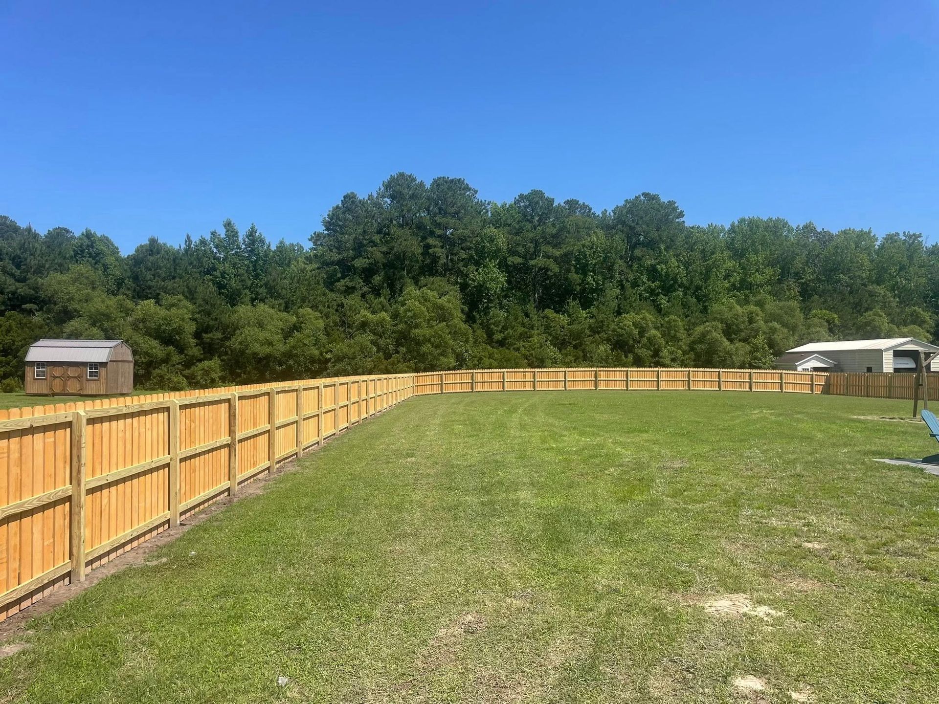 Wooden fence encloses a grassy backyard; trees and blue sky in the background.