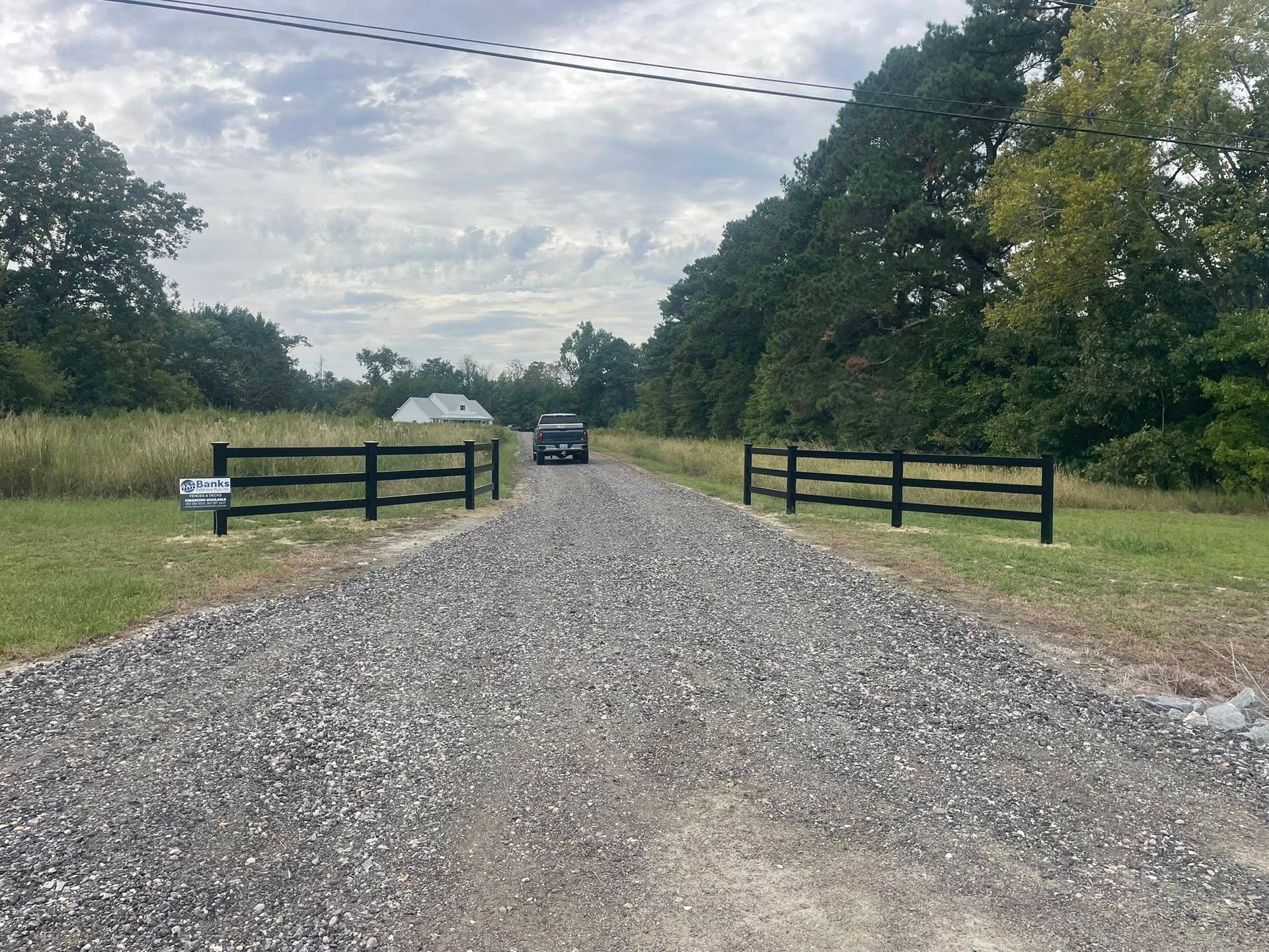 Gravel driveway leading to a car, flanked by black fences, under a cloudy sky.