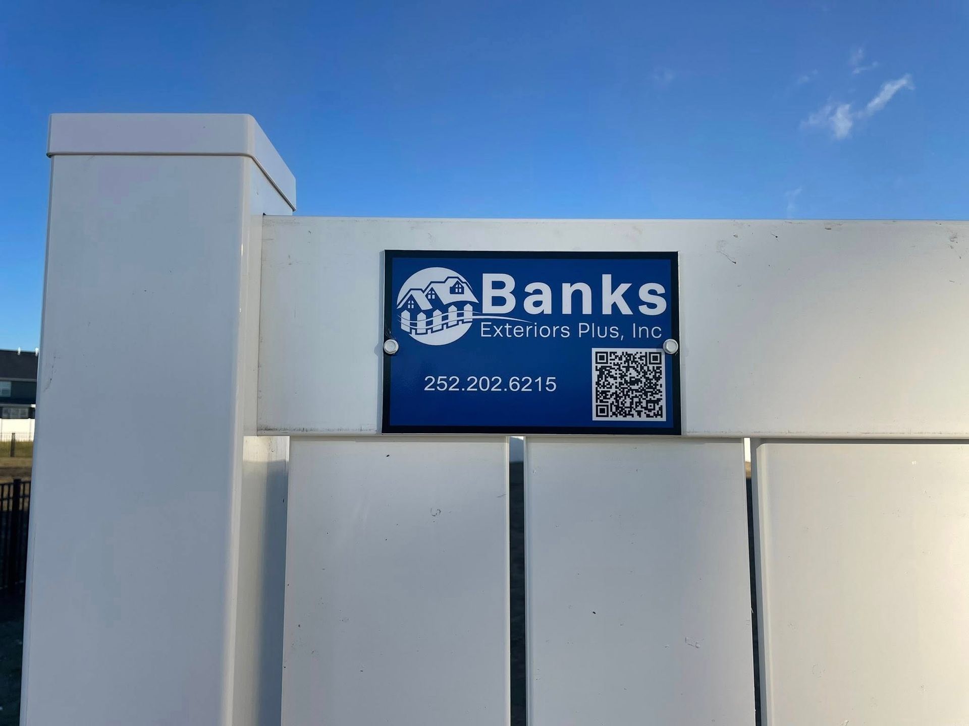 Sign for Banks Collision Plus, Inc. mounted on a white fence with a QR code and phone number, against a blue sky.