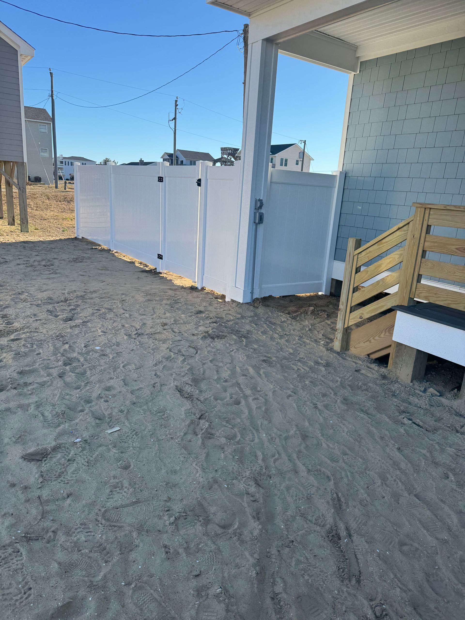 White fence and gate next to a house with wooden steps, set on sand, bright sky.