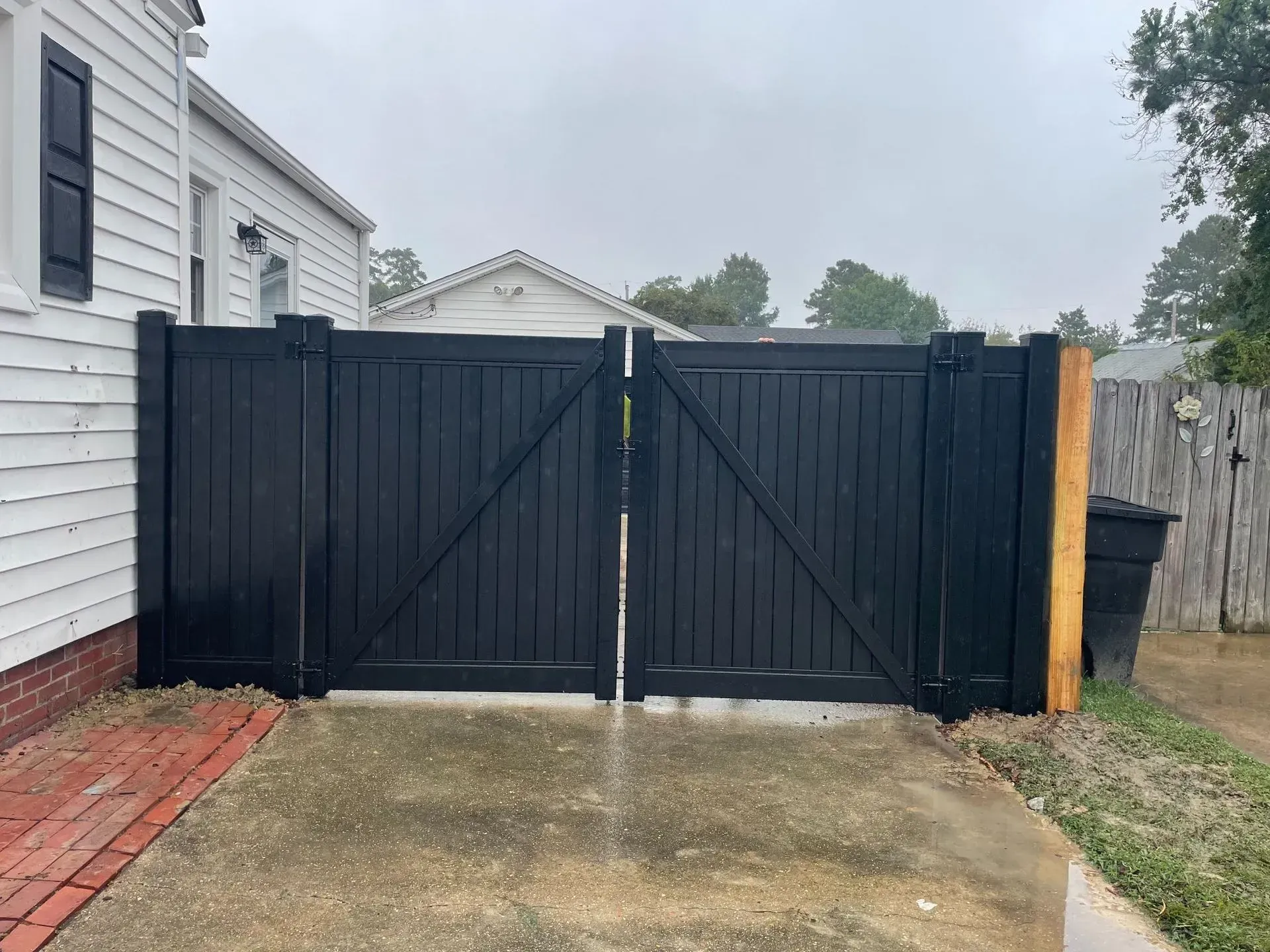 Black wooden gate in front of a white house, on a concrete surface. Adjacent to a fence.