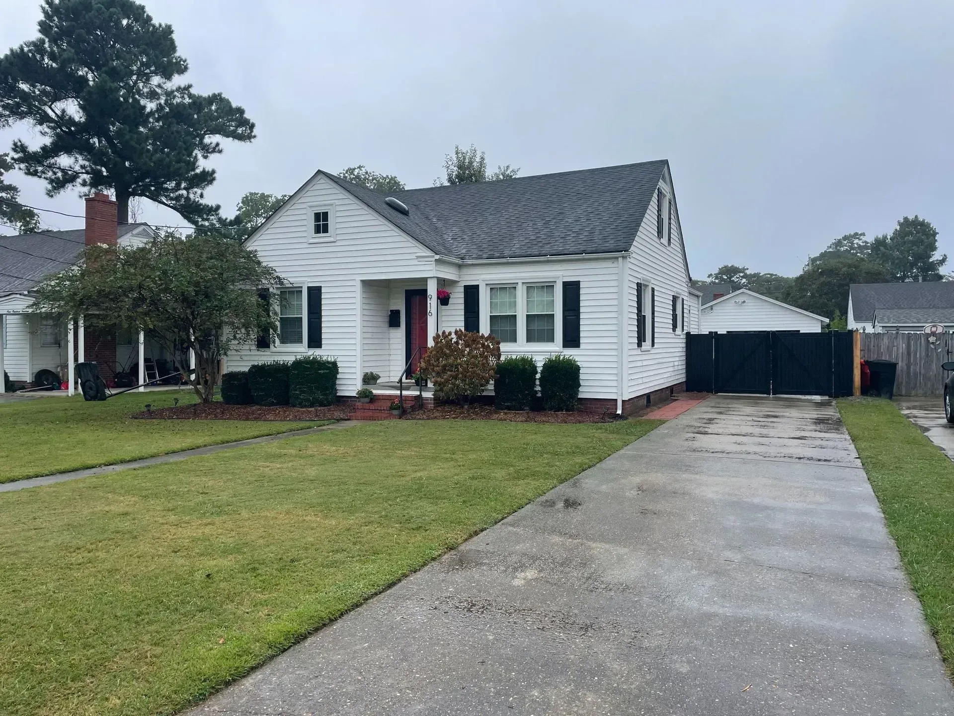 White house with black shutters, red door, and driveway on a cloudy day. White house with black shutters, red door, and driveway on a cloudy day.