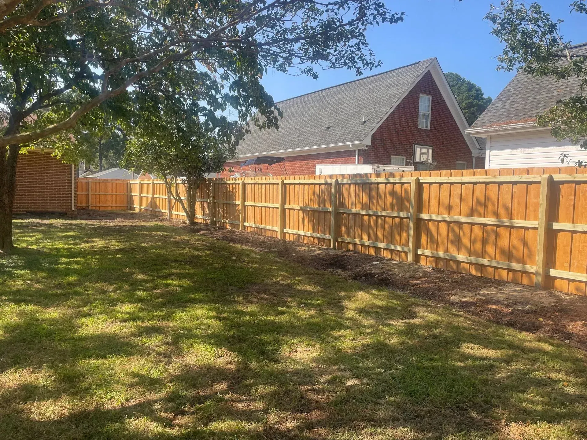 Wooden fence in a backyard with a house in the background. Green grass and a tree are in the foreground.
