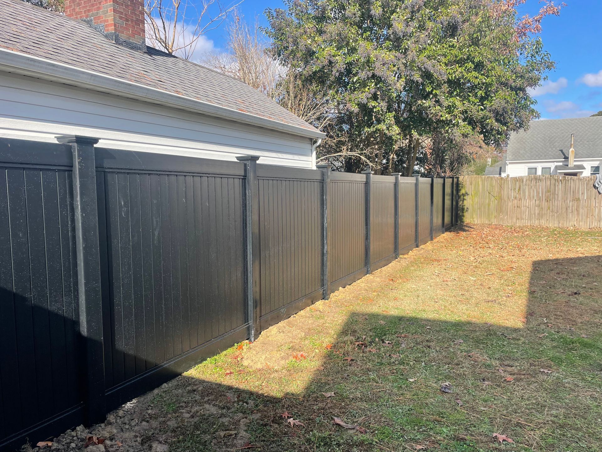 Black vinyl fence bordering a yard with brown leaves, alongside a house with a brick chimney.
