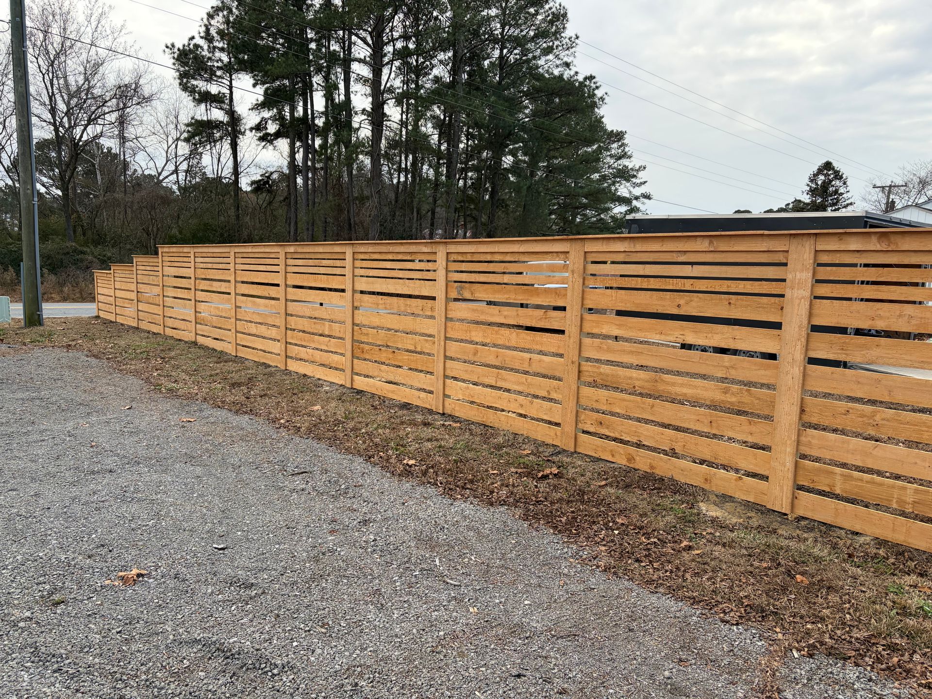 Wooden horizontal slat fence along a gravel area with trees in the background.