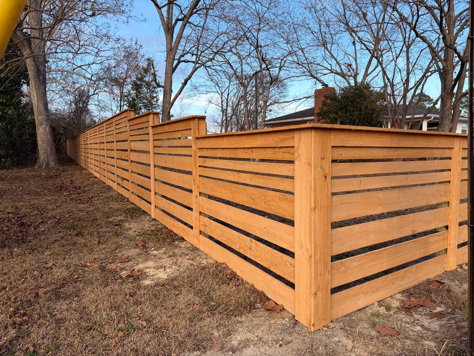 Wooden horizontal slat fence in a yard with bare trees and a blue sky.