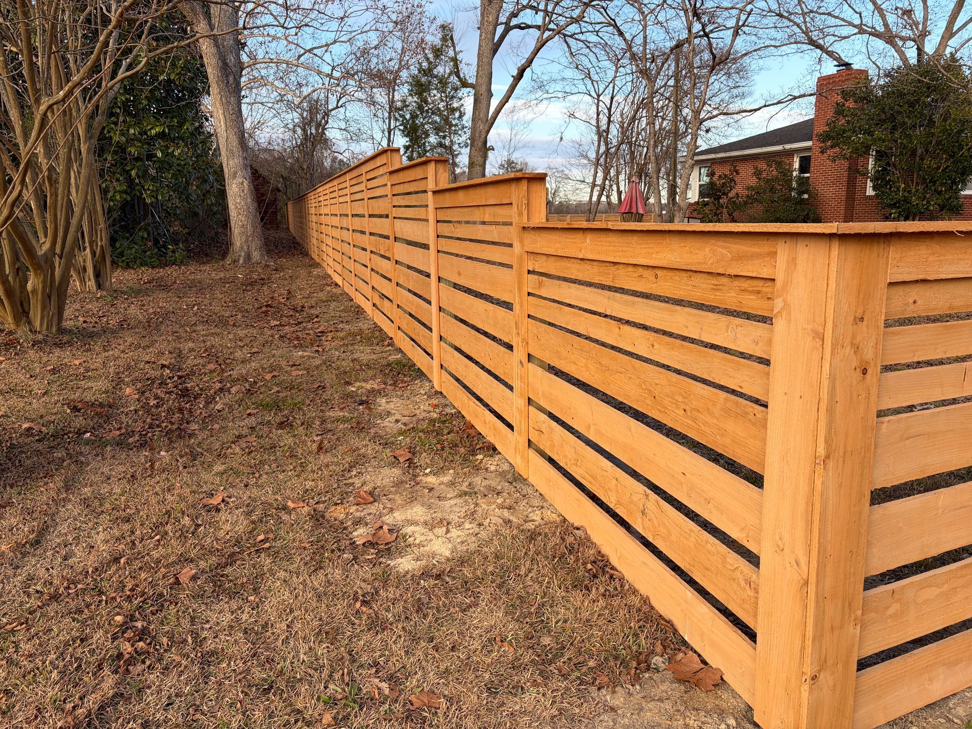 Wooden horizontal slat fence in a yard with bare trees and brown leaves.