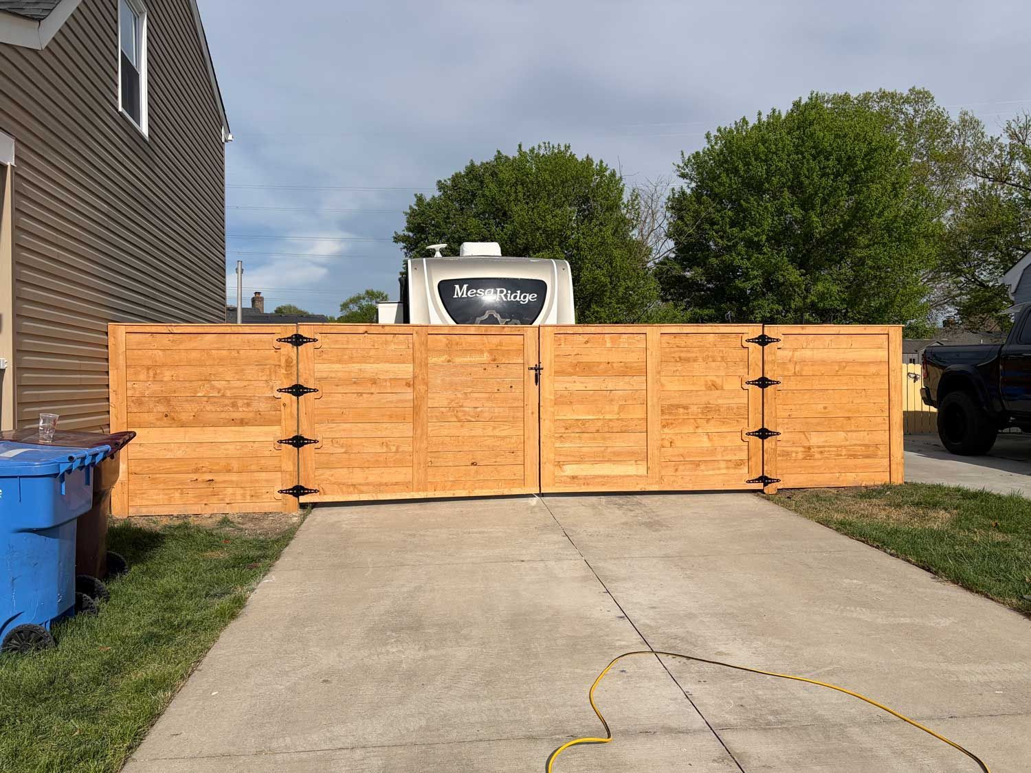 A wooden gate installed across a residential concrete driveway, with a blue bin on the left and a camper in the background.