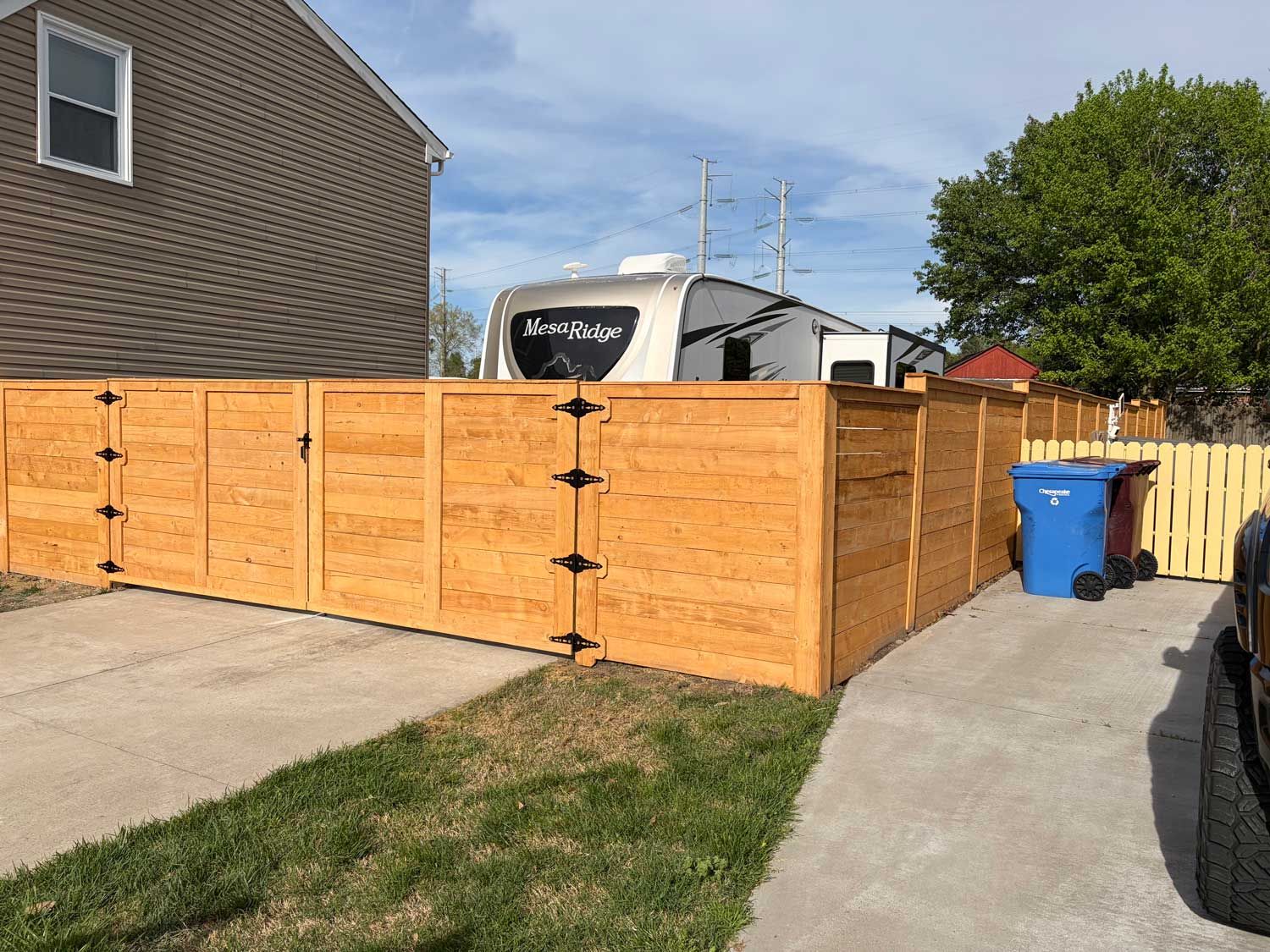 A tall wooden privacy fence wraps around a corner on a concrete driveway, with a camper parked behind it.