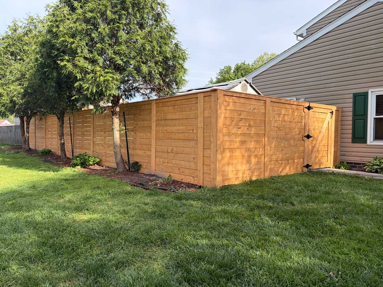 A tall, stained wooden privacy fence with a gate, installed beside a tan house and green trees on a sunny lawn.