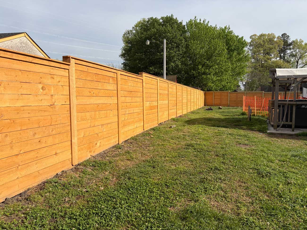 A long, horizontal wooden privacy fence runs along a green, grassy yard under a bright, partly cloudy sky.