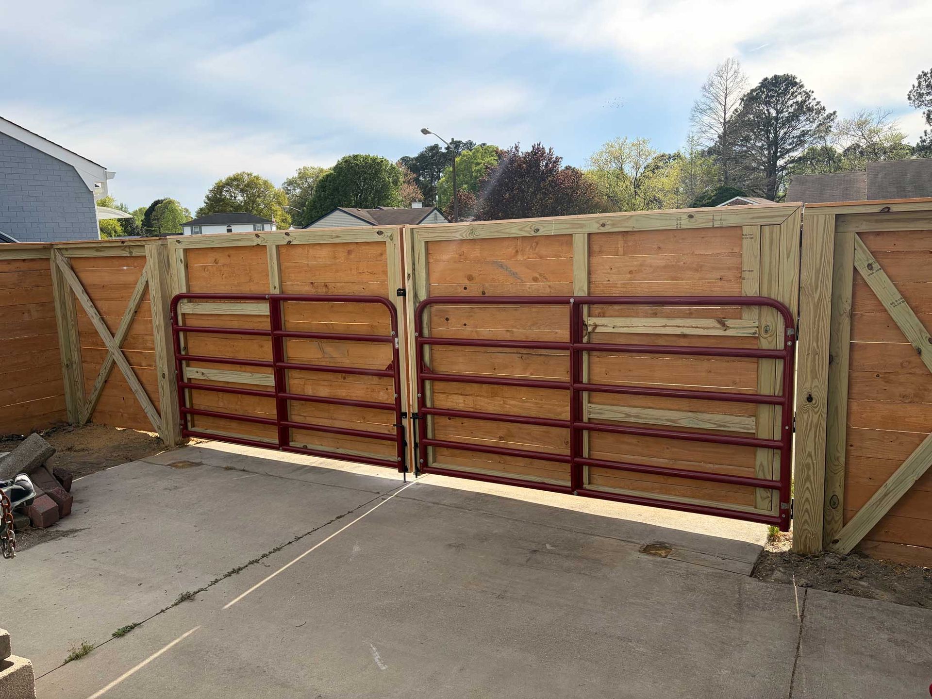 A pair of dark red metal farm gates installed within a wooden fence enclosure on a concrete driveway.