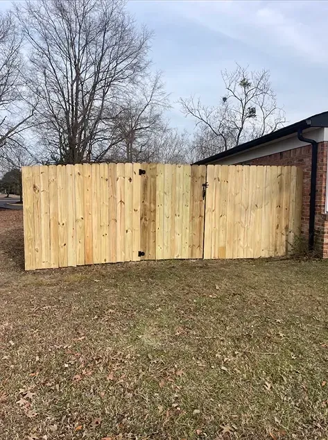Wooden fence with gate, bordering a grassy yard next to a brick building, under a cloudy sky.
