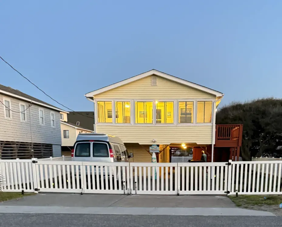 Yellow house with white fence and parked van; lights on in the windows.