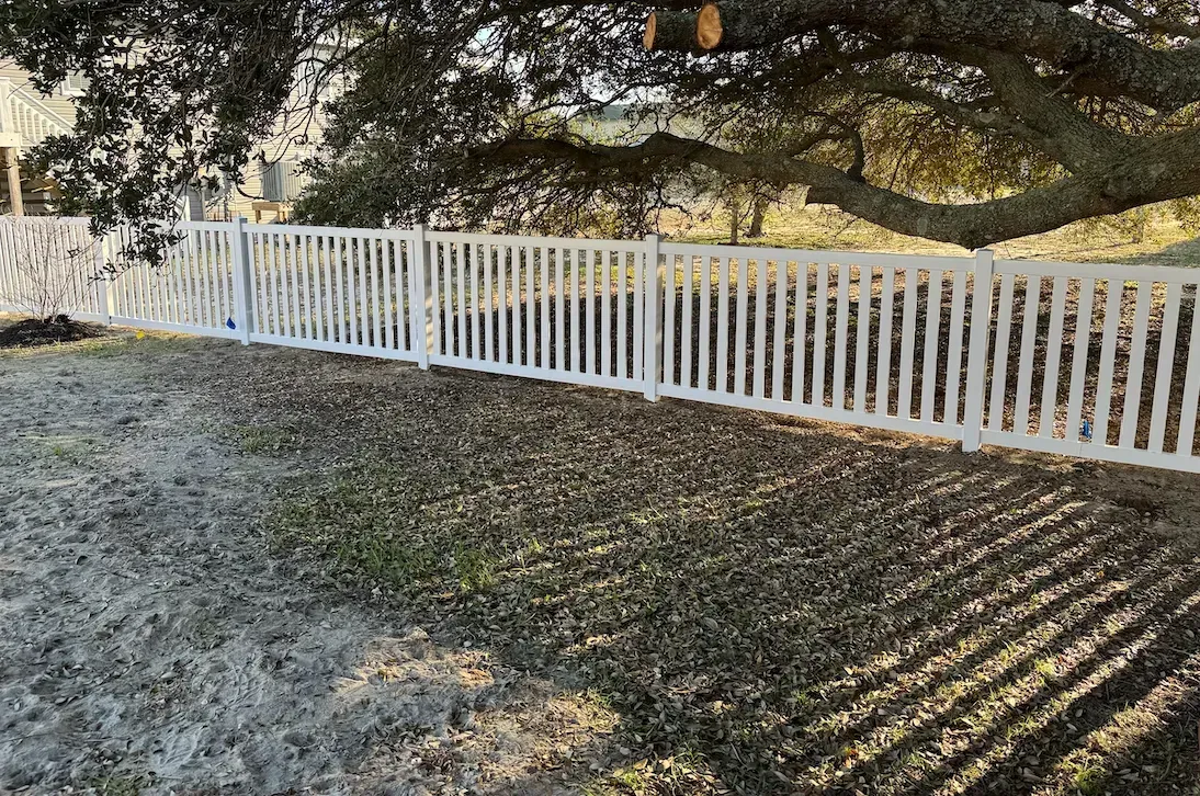 White picket fence under a large tree, in a grassy yard, casting shadows.