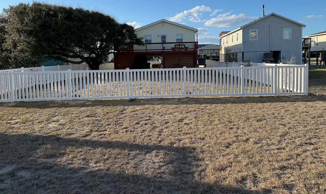 White picket fence surrounding a yard with two-story houses and a tree in the background.