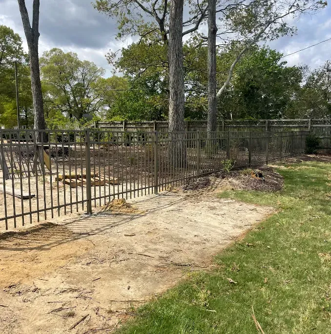 Metal fence surrounding a sandy area, trees and grass in the background.