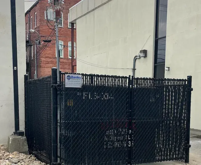 Black chain-link fence encloses an outdoor space; a brick building and a tan wall are in the background.