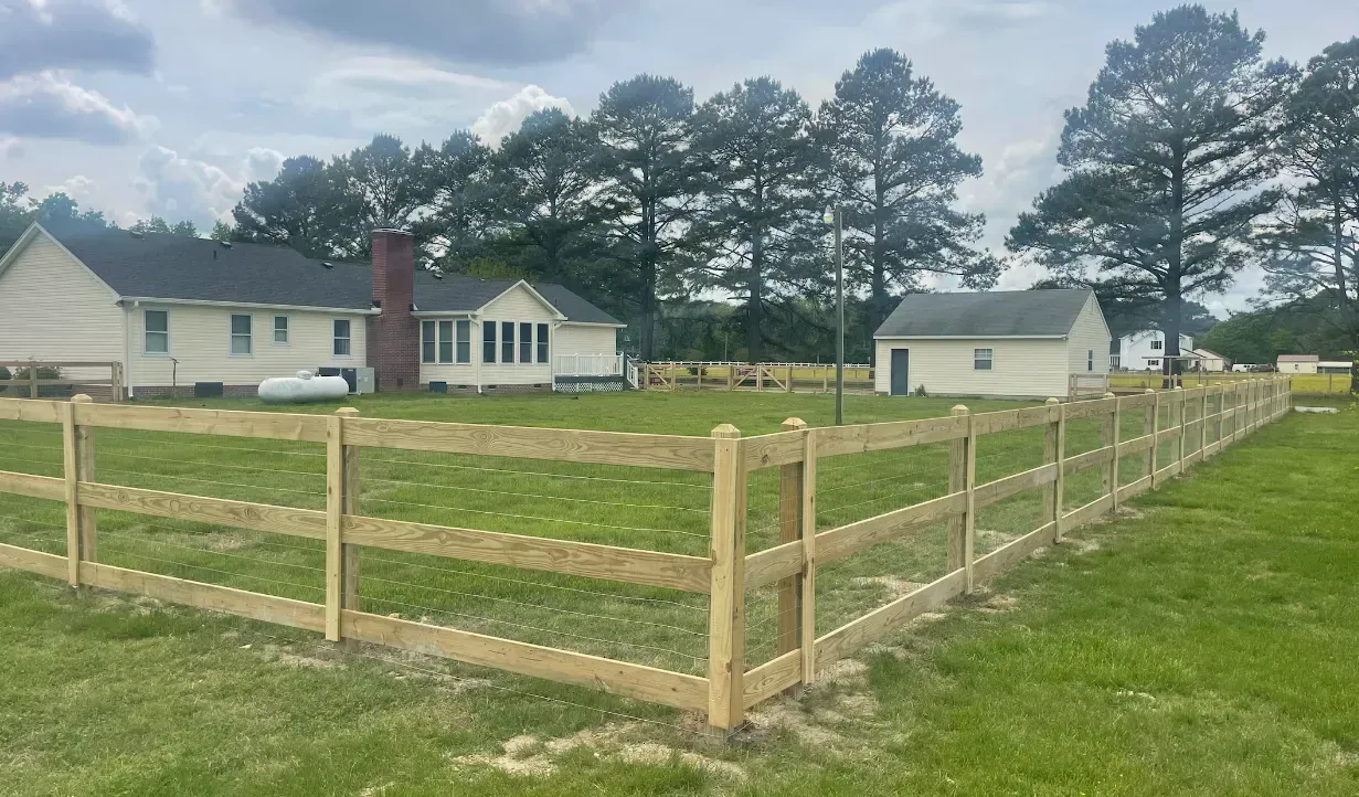 Wooden fence surrounding a grassy yard with a house and outbuildings in the background. Overcast sky.