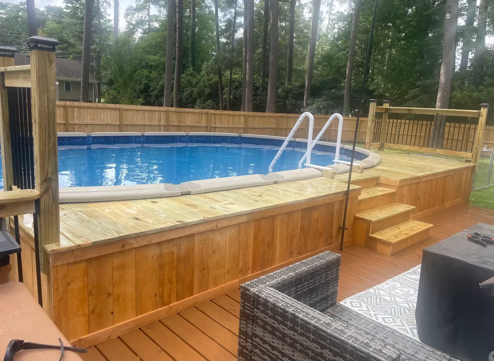 Above-ground pool with wooden deck and stairs, fenced backyard.