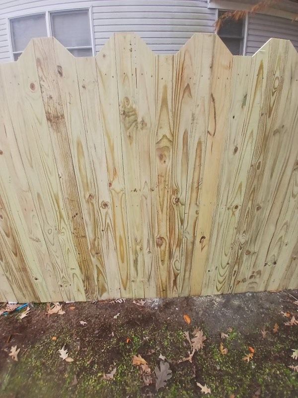 Wooden fence with scalloped top, built next to a building with windows. Ground with leaves in front.