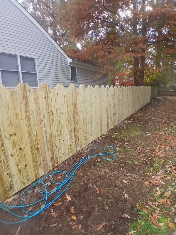 Wooden fence in front of a gray house, blue hose on the ground, trees in the background.