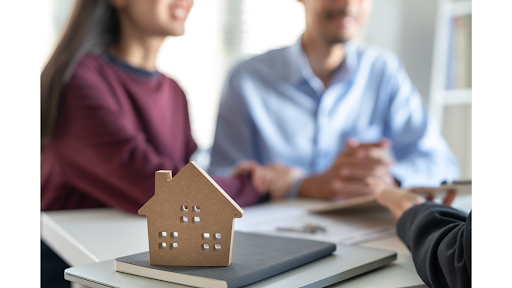 A small wooden house model sits on a table while a couple is seated smiling while interacting with a real estate agent.