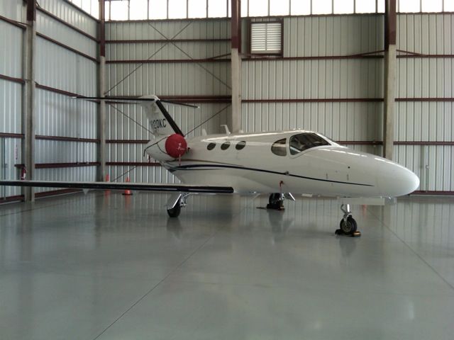Small white jet airplane parked inside a hangar with a red engine cover and hangar doors visible