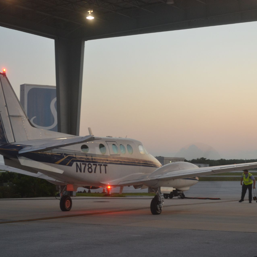 Small twin-engine airplane parked on a tarmac at dusk, with a person in a reflective vest nearby.