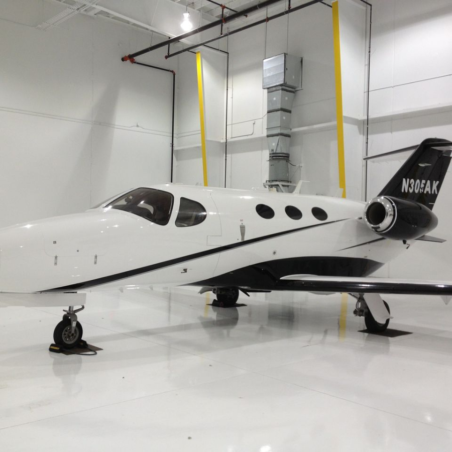 Small white private jet parked in a bright hangar, with black tail and yellow support straps.