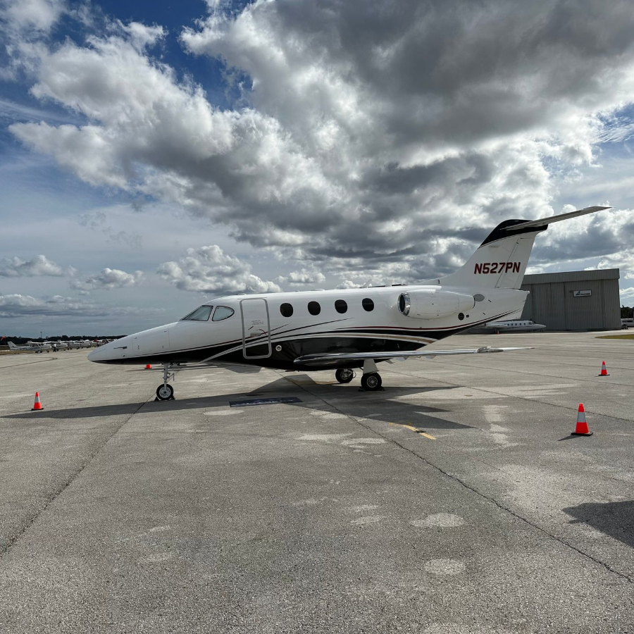 Small white private jet on a tarmac under cloudy sky, with orange cones nearby.