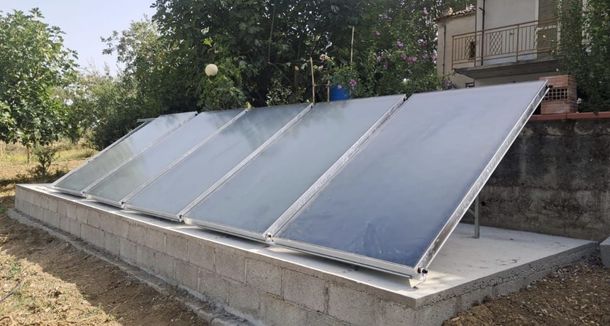 Una fila di pannelli solari inclinati per il riscaldamento dell'acqua su un tetto accanto a una casa e agli alberi.