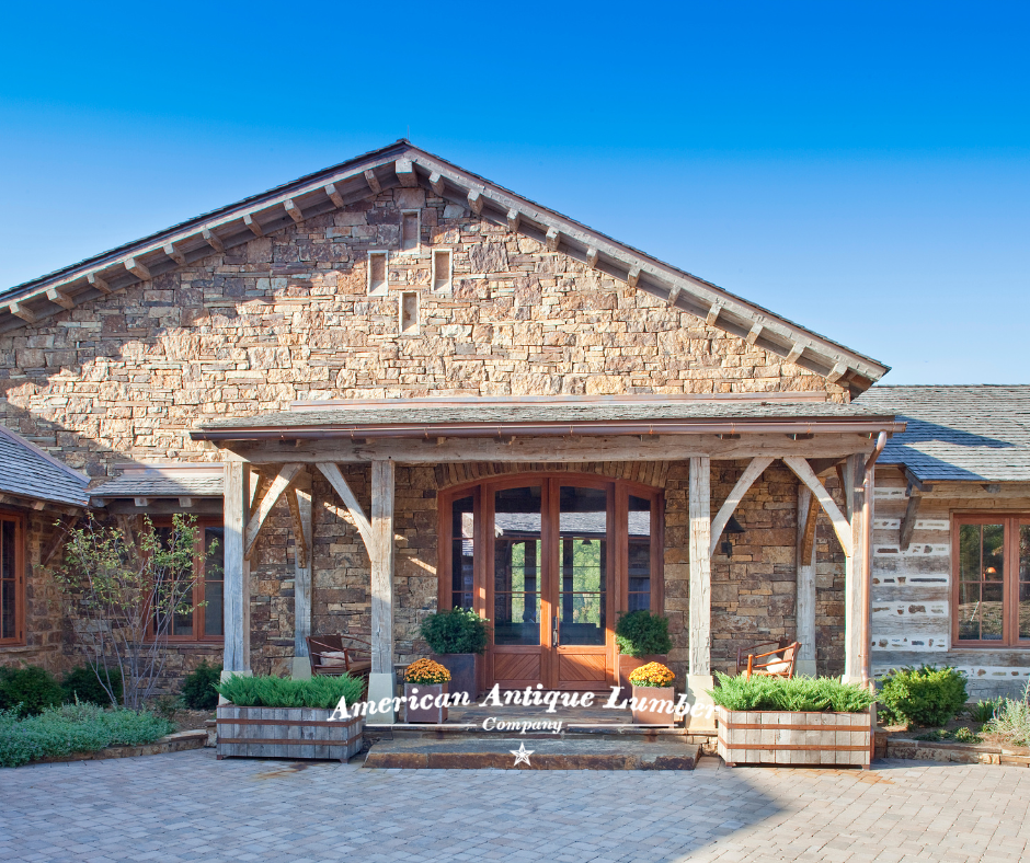 Exterior stone wall with a wooden and glass front door and reclaimed hand hewn ornamental porch posts and beams with green plants in planters. 