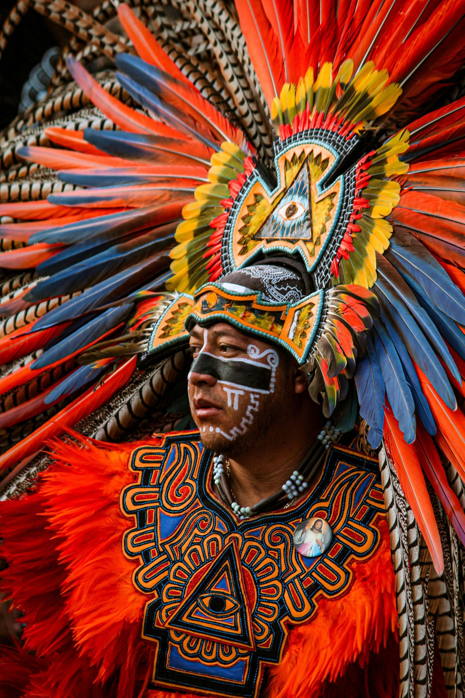 Man in colorful Aztec ceremonial headdress and clothing, dark face paint, feathers, and elaborate patterns.