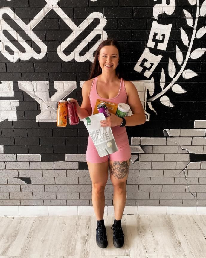 A Woman is Standing in Front of a Brick Wall Holding Health Products and Supplements — The Factory Gym in Taree, NSW