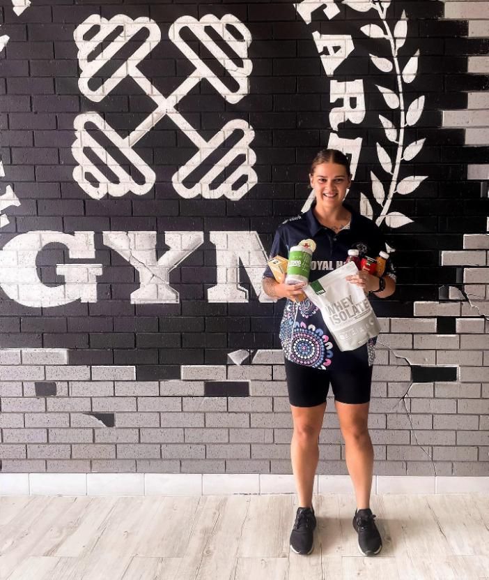 A Woman is Standing in Front of a Gym Wall Holding Health Products and Supplements — The Factory Gym in Taree, NSW