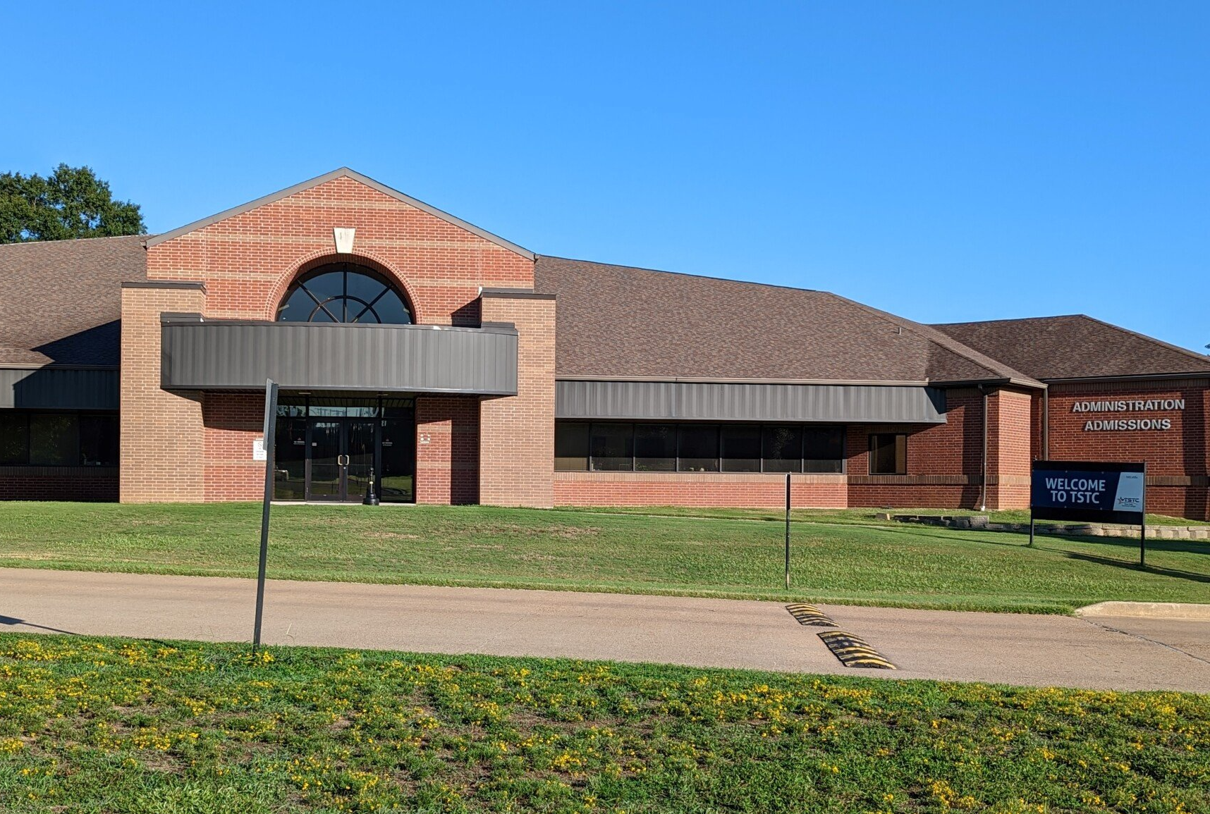 A large brick building with a metal roof is surrounded by grass and flowers.