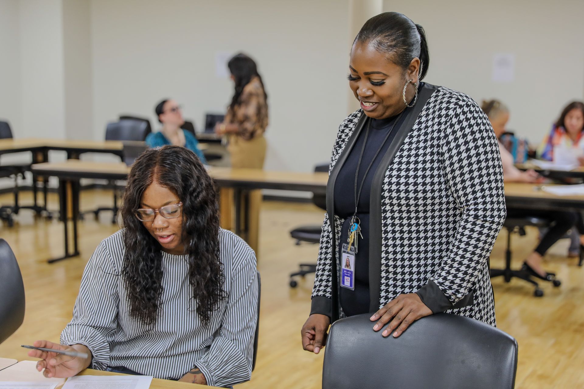 A woman is standing next to a woman sitting at a desk in a classroom.