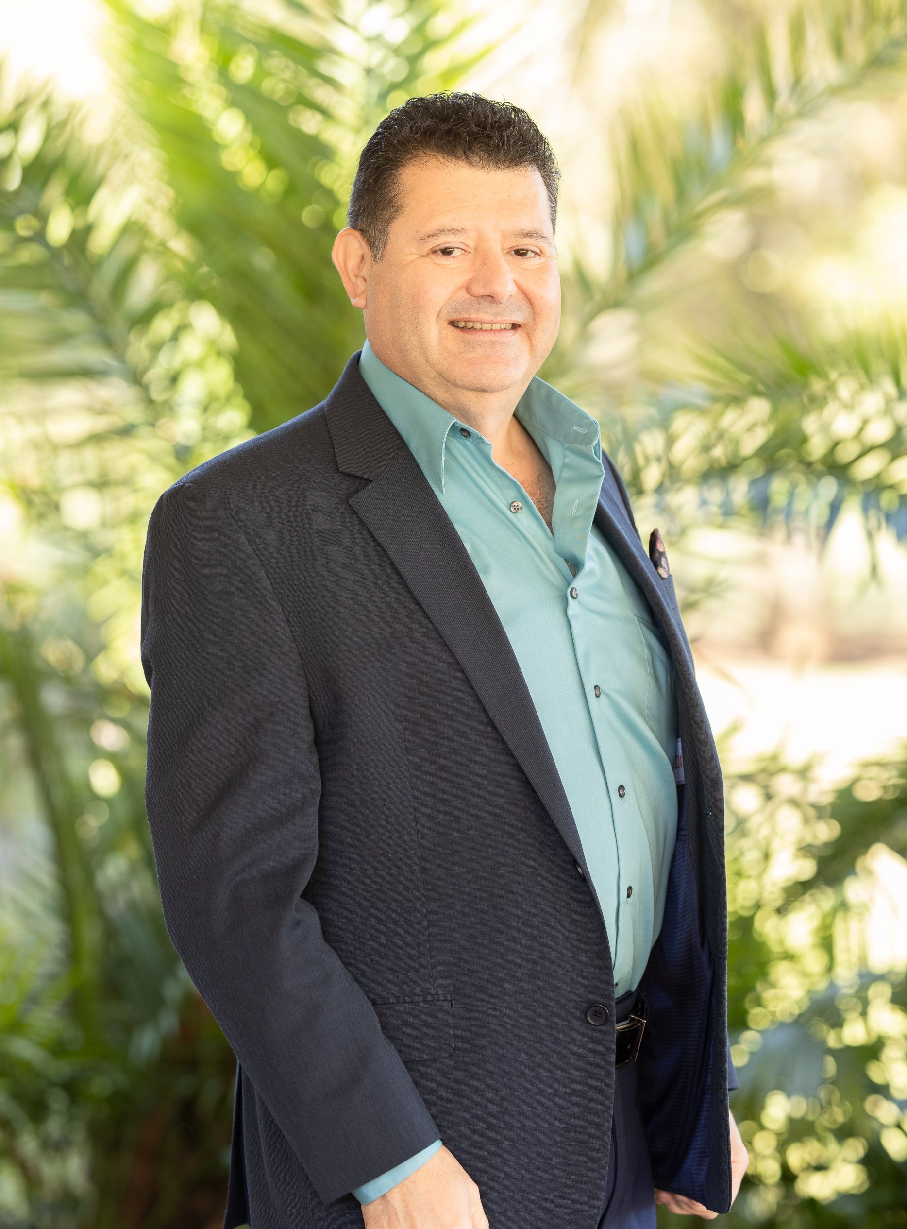 A man in a suit and green shirt is standing in front of a palm tree.