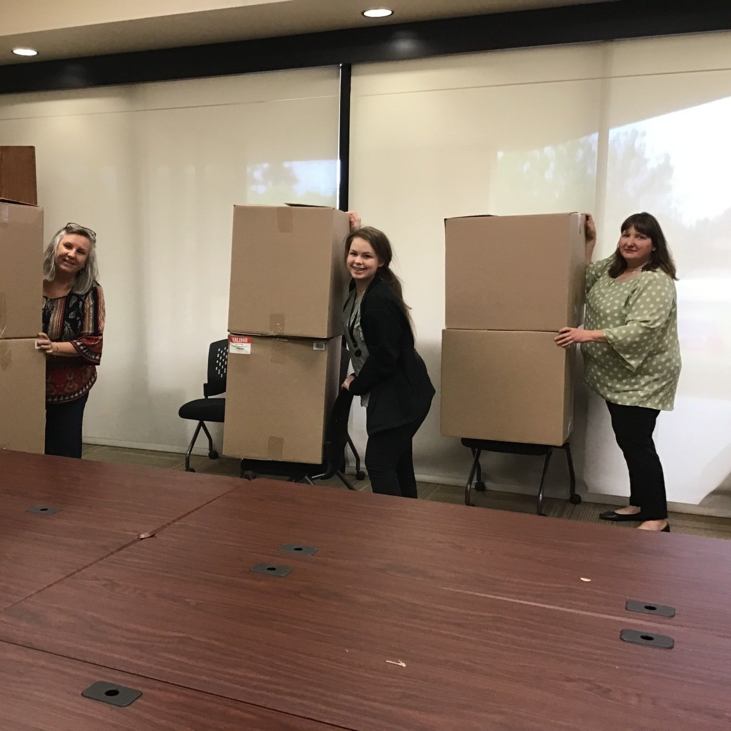 Three women are standing next to each other holding cardboard boxes in a room.
