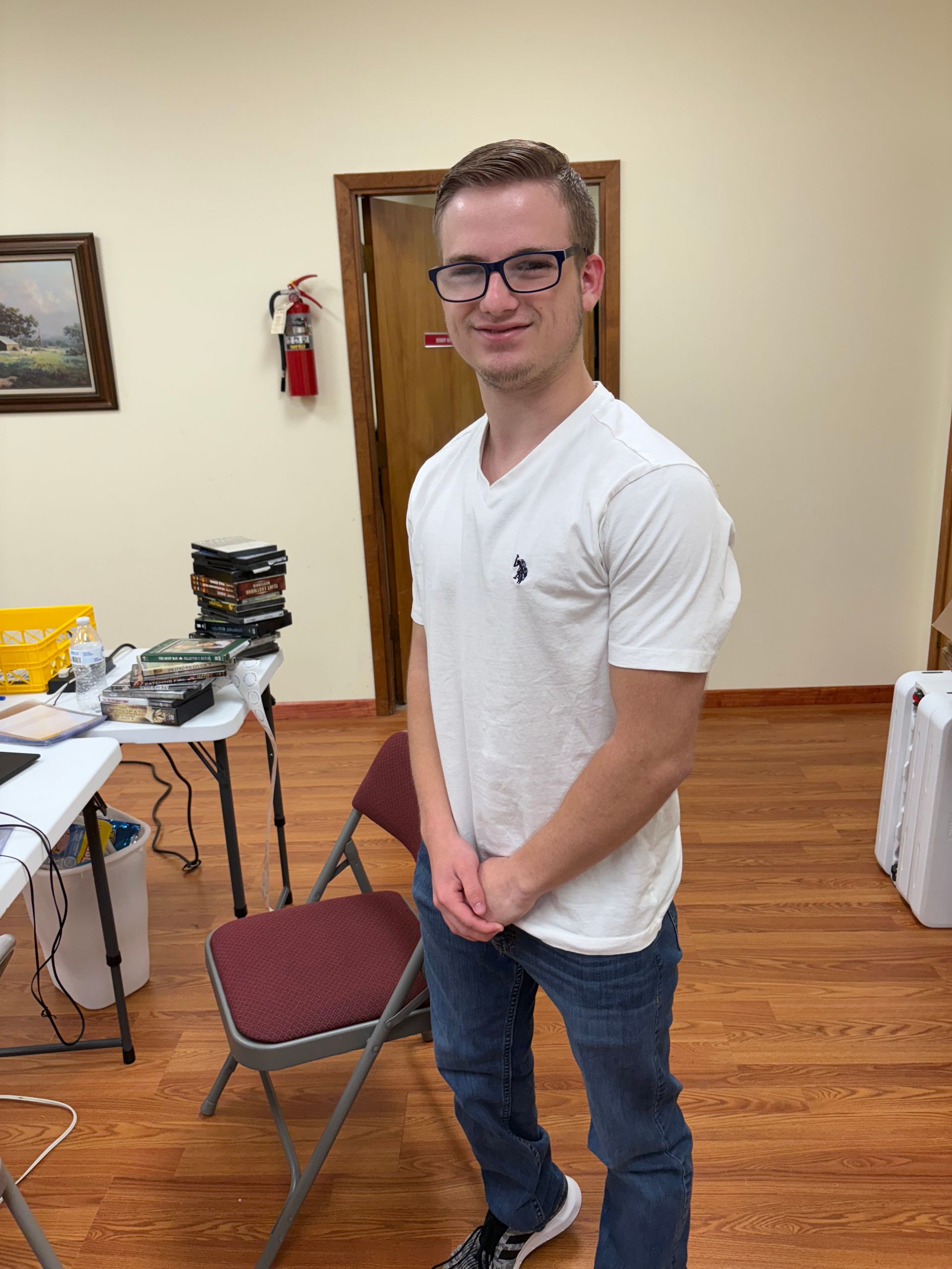 Man in glasses and white shirt stands in a room with a table and chairs.