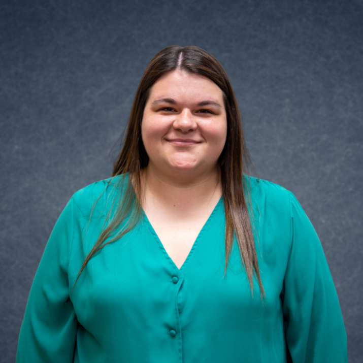 Smiling person in a teal blouse standing against a dark blue backdrop