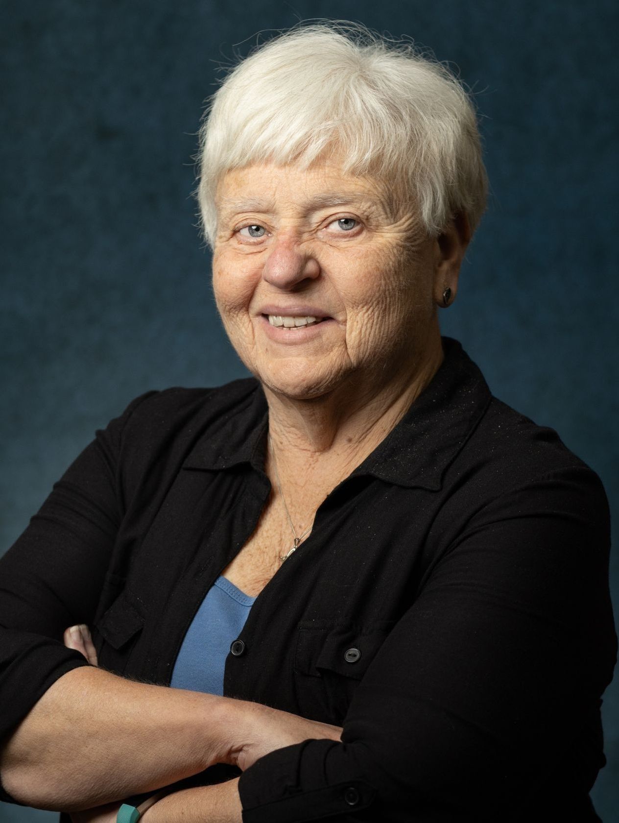 A woman with white hair is wearing a black shirt and smiling with her arms crossed.