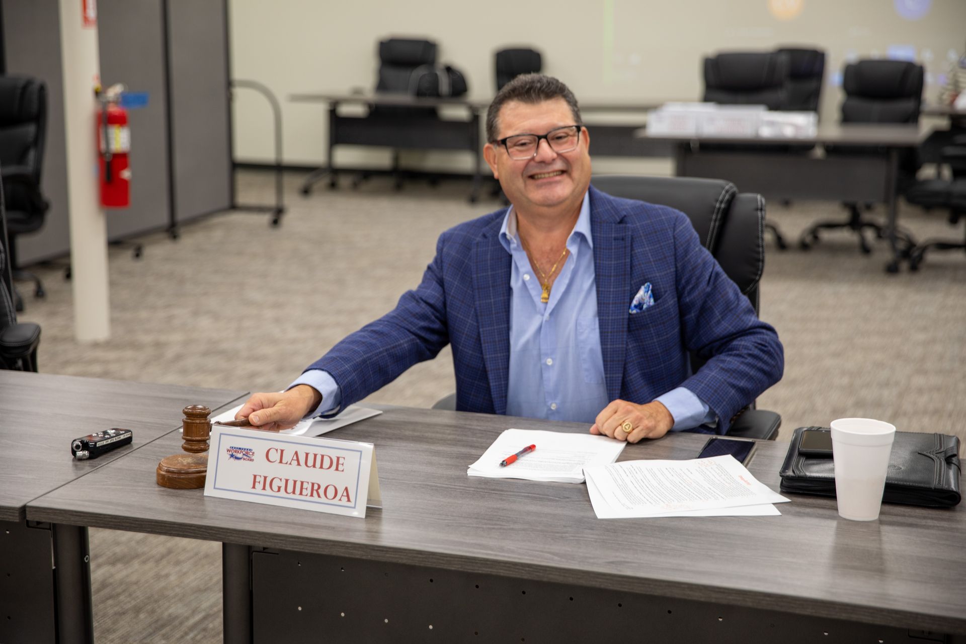 Man in blue blazer smiles, seated at a table with a nameplate (