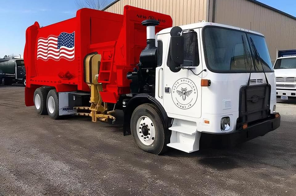 White and red garbage truck with an American flag on the side, parked outside a building.